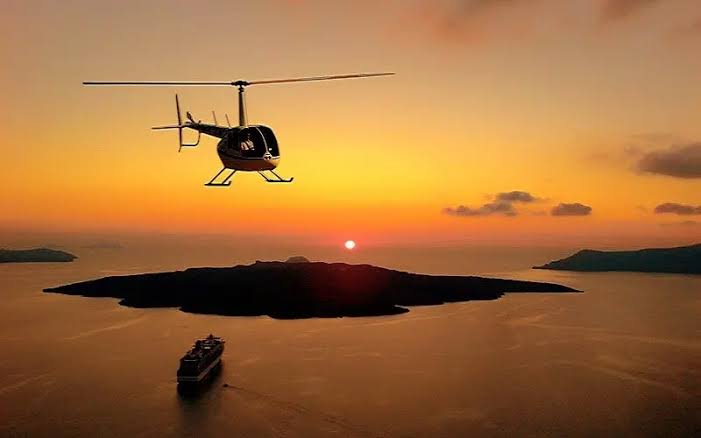 An Ariston Aviation private helicopter flying gracefully into a golden sunset over the calm waters of the Aegean Sea